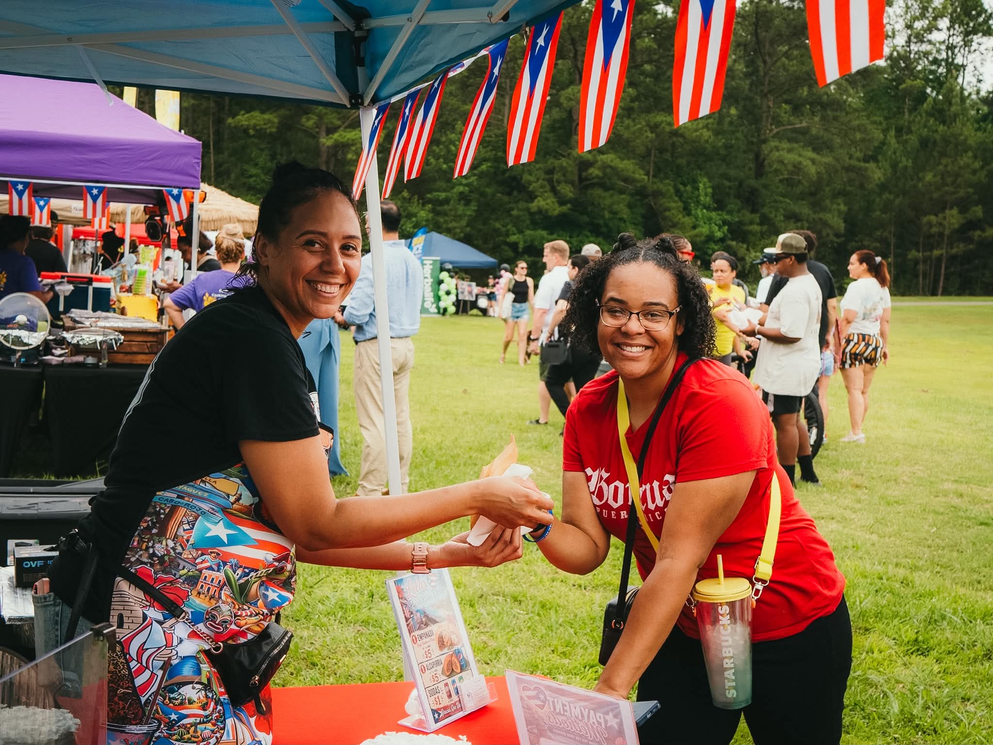 Two women smile from a food booth beneath Puerto Rican flags at the festival.